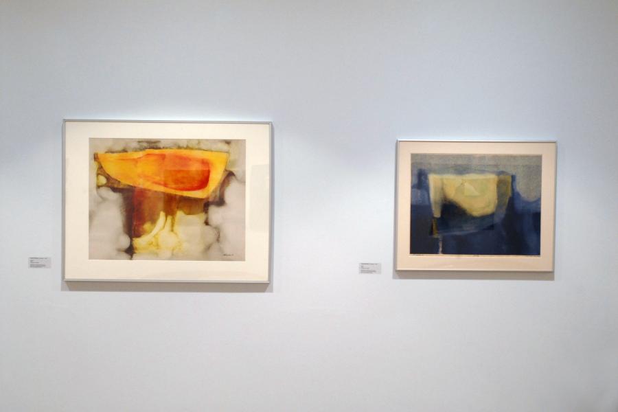 Two abstract graphics on a wall. The left one comprises an orange, yellow, and brown form on a white background. The right one comprises a yellow form on a blue background and a grey section on top.