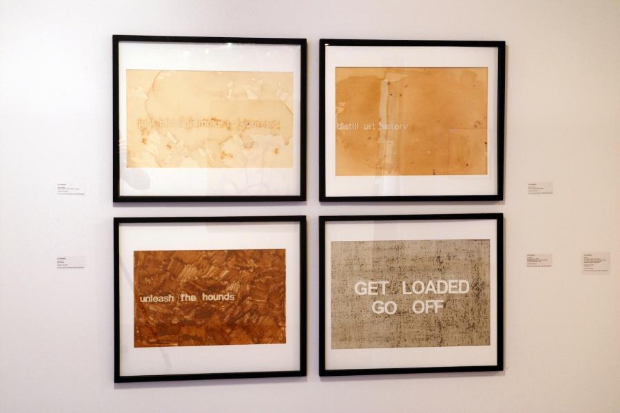 Four artworks on a wall, comprising sans-serif text on coloured backgrounds. Clockwise from the top left, they read “just blowing smoke up your ass” in lowercase light beige text on a stained light beige background, “distill art history” in lowercase light beige text on a light brown background with some dark spots, “unleash the hounds” in light red text on a dark red streaked background, and “get loaded go off” in uppercase white text on a dark beige background covered in dark spots.