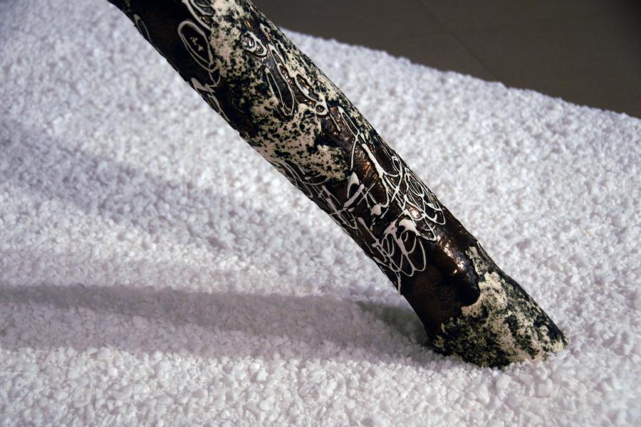 Close-up on the base of a sculpture depicting a birch tree trunk in the snow. The sculpture is mostly black, covered in messy white lines and textures. It is surrounded by a bed of small white rocks.