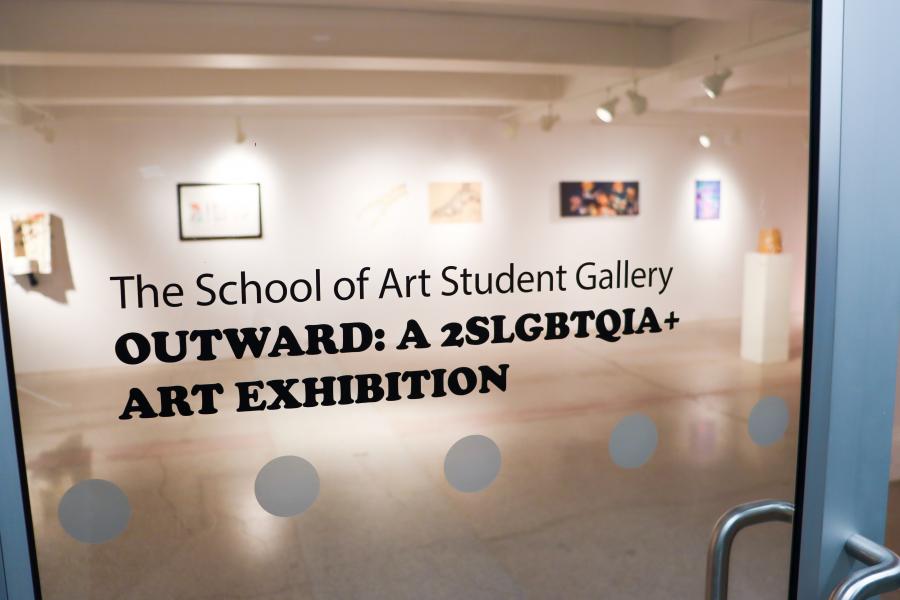 Glass gallery entrance with vinyl text reading “OUTWARD: A 2SLGBTQIA+ Art Exhibition,” with artworks visible inside the gallery.
