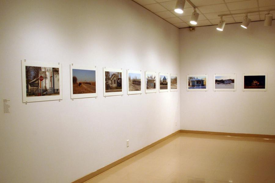 Ten street photographs hung in a line along the corner of two walls. a street sign reading “logan ave” in front of a white church, a road crossing over a creek surrounded by dead plants, a white house, a tree-lined dirt path, a blue house, a yellow house, an orange corner store, another white house, a fallen letter board, and a Christmas light display at night.