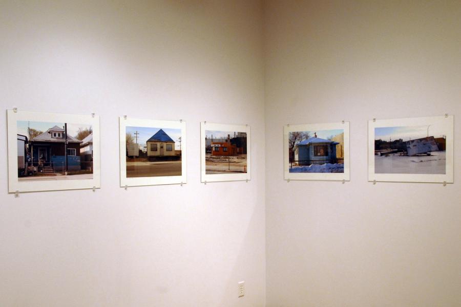 Five street photographs hung in a line along the corner of two walls. They feature a blue house, a yellow house, an orange corner store, a white house, and a fallen letter board.