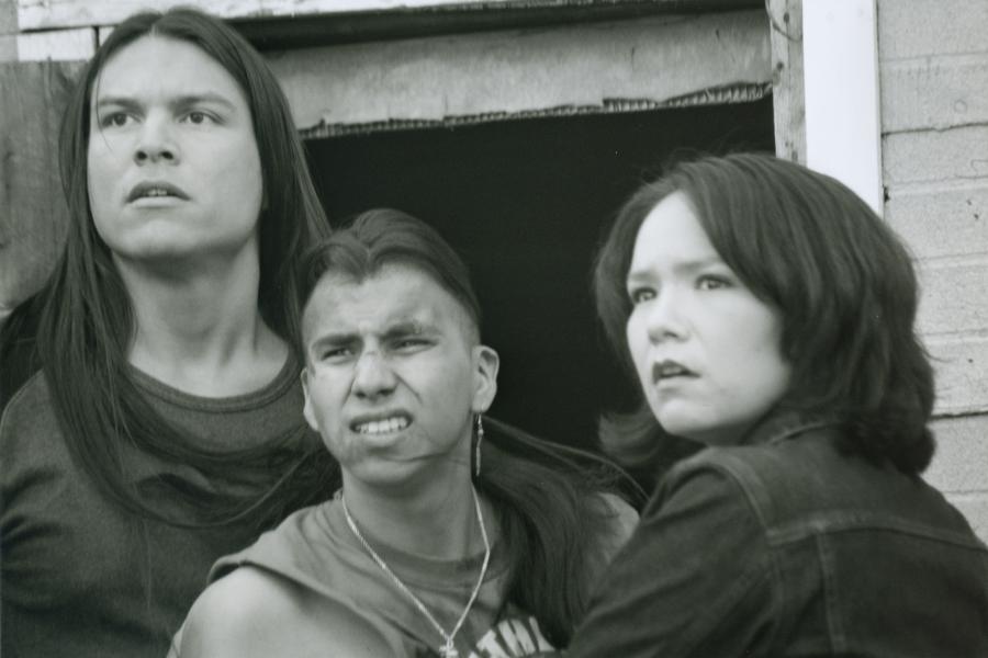 Black-and-white film still showing three young people standing close together outside a weathered building, all looking off-frame with concerned expressions.