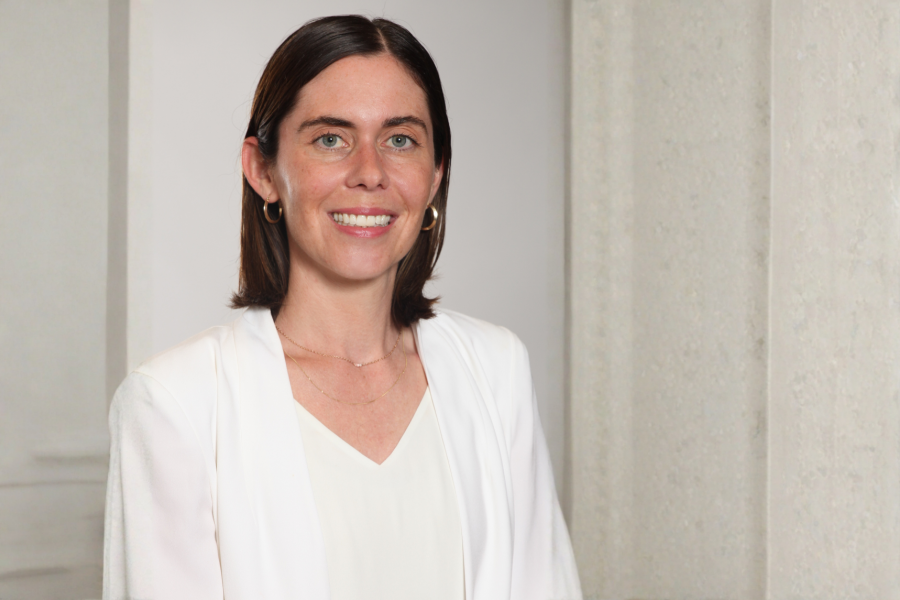 Portrait of Breanna Lawrence with shoulder-length dark brown hair smiling at the camera, wearing a white blazer over a light top with small gold hoop earrings and layered necklaces
