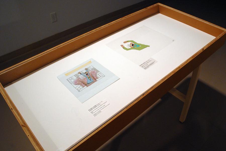 Two stills of cartoon men in coloured sweaters and bowties screaming with their arms raised, displayed in a wood case. The left one depicts its subject sitting in a pink chair at a table, while the right’s subject is drawn on a transparent field.