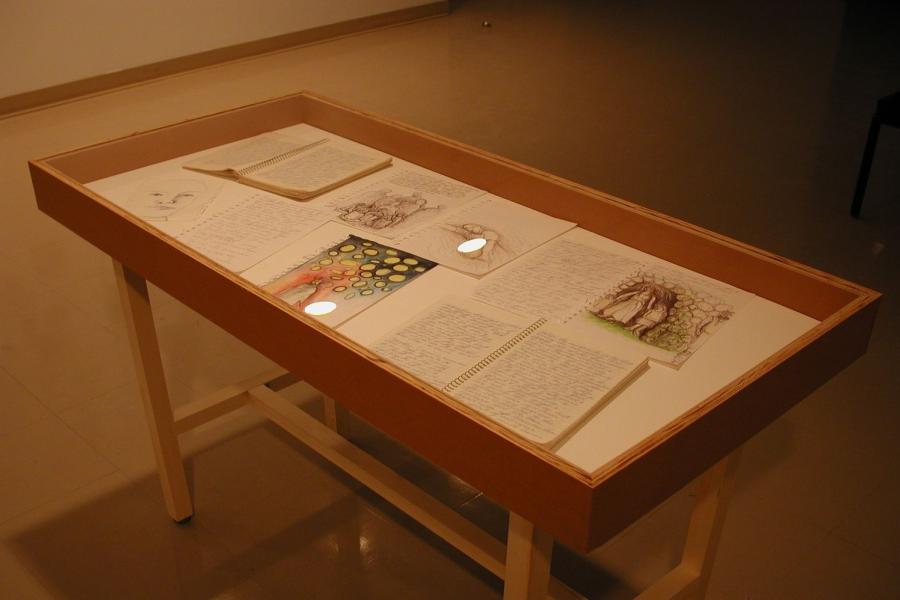 A display case showing six open notebooks of handwriting and sketches. They are sketches of a face, a figure’s silhouette on a bright red and black background with yellow circles above them, a crowd of figures’ silhouettes, a figure lying down, and a woman with two children in a stone archway and another person standing outside.