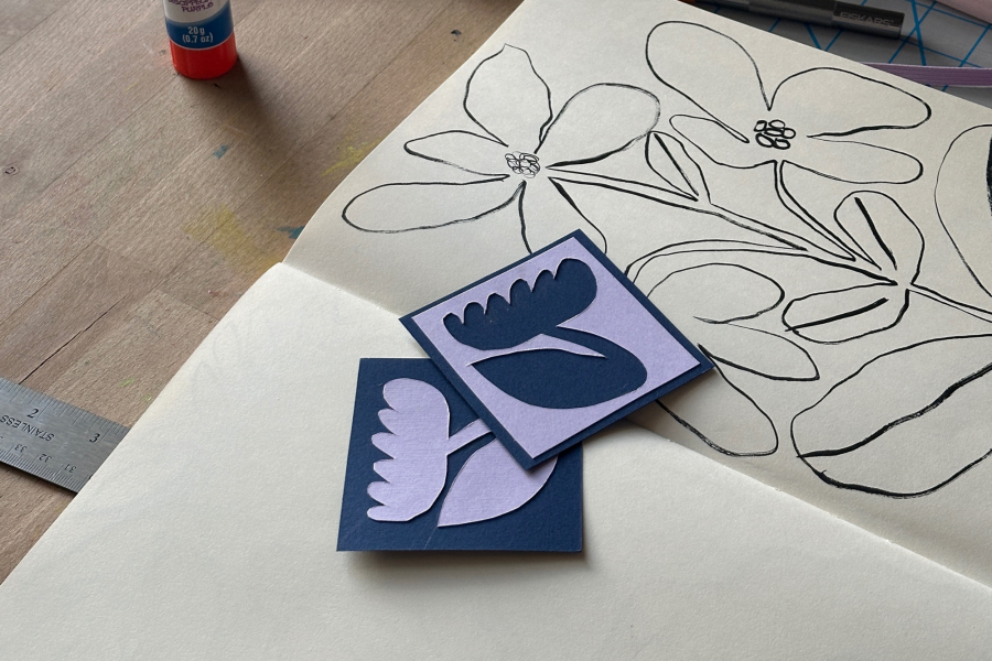 Sketchbook open to a black ink drawing of flowers, with layered paper cut-out shapes in purple and blue resting on top, arranged on a wooden table.
