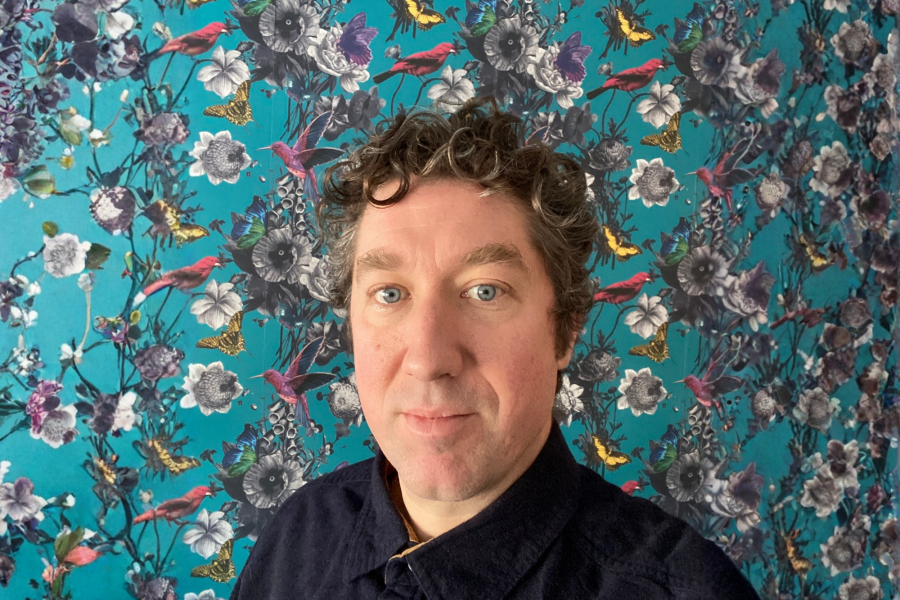 Portrait of David Peters with short curly hair standing in front of a teal patterned background featuring flowers, birds, and butterflies.