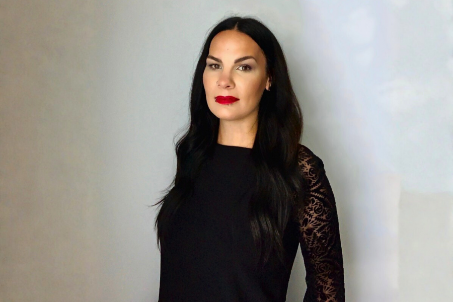 Studio portrait of Renata Truelove with long dark hair standing against a light background, wearing a black top with lace sleeves and red lipstick, looking directly at the camera with a composed expression.