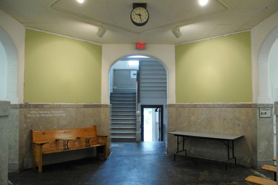 The rotunda outside of the gallery, fitted with large rough-edged green rectangles occupying the upper halves of the corner walls, above the stone dados. Wall text reads “Jeffery Spalding: Neues Bild (Rotunda, Winnipeg) 1976/200” under the one on the left. A wooden bench sits against the wall on the left, and a folding table sits beneath the wall on the right, and a staircase is visible behind the archway in the centre of the image.