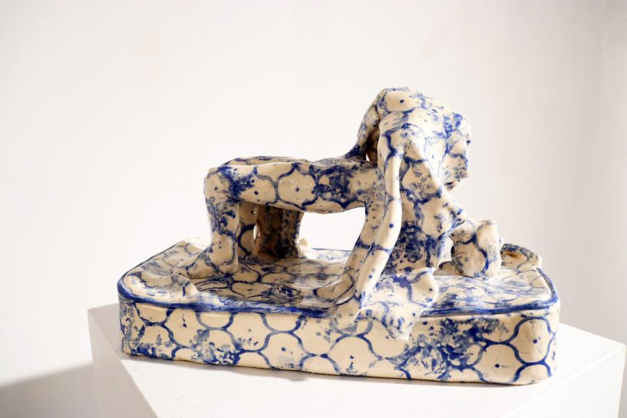 Ceramic sculpture of an arched human figure on a rectangular base, glazed in cream with blue marbled patterns, displayed on a white pedestal.