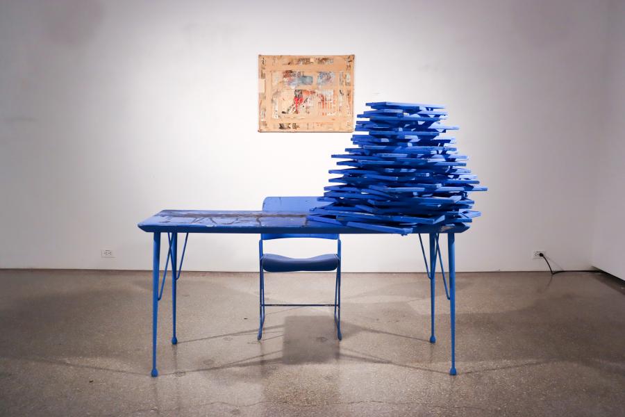Blue table and chair in a gallery with a tall stack of blue panels on the tabletop and a framed abstract artwork hanging on the wall behind.