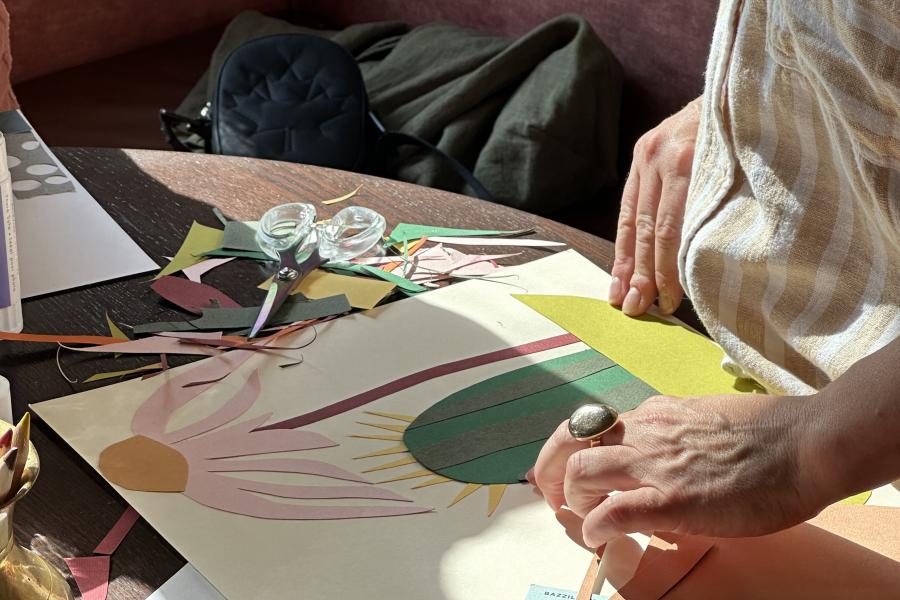 Sketchbook open to a black ink drawing of flowers, with layered paper cut-out shapes in purple and blue resting on top, arranged on a wooden table.
