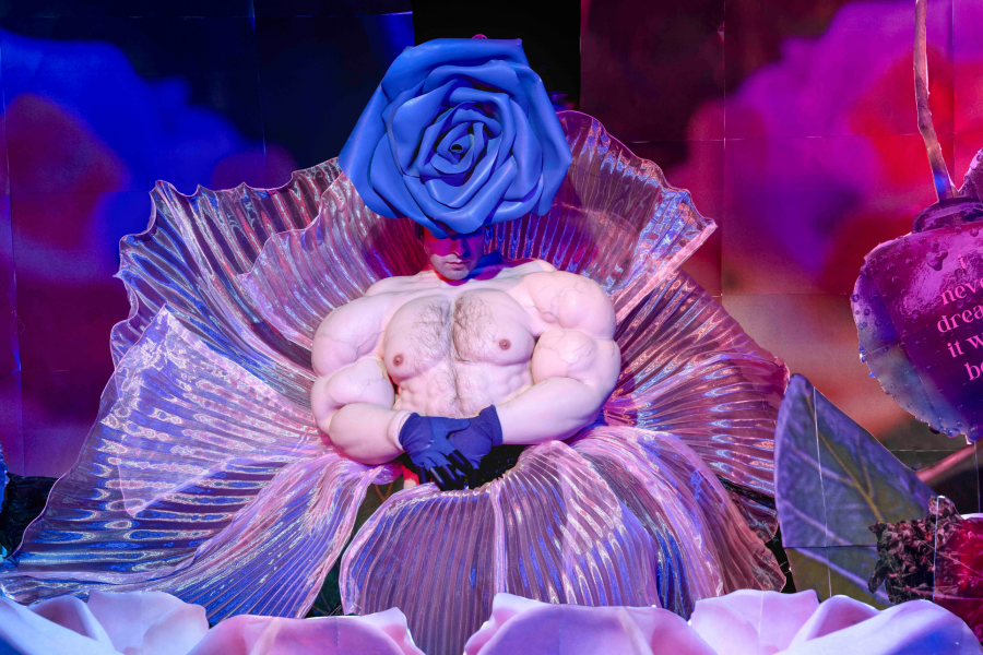 A staged portrait of a performer seated inside an iridescent flower-like form, wearing exaggerated muscular prosthetics and a large blue rose headpiece, surrounded by glowing pink and blue floral imagery and oversized cherries.