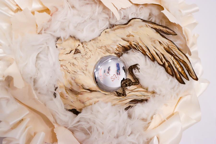 Close-up of a mixed-media wall sculpture featuring painted bird imagery surrounded by white feathers and curled paper elements, with a reflective circular mirror at the centre reading “need safe.”