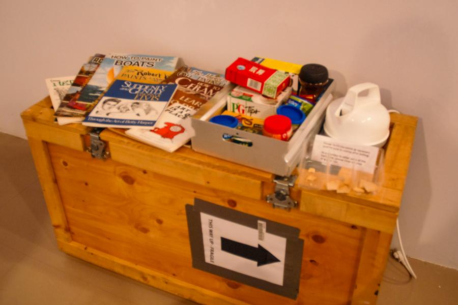 Detail of Dick Averns' installation featuring a wooden shipping crate topped with books, packaged food, and everyday household items arranged as part of the artwork.