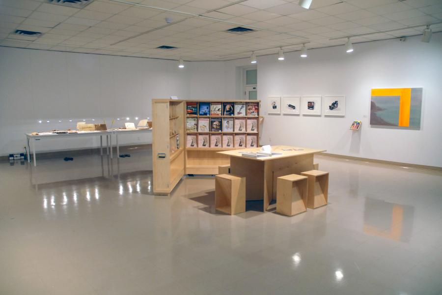 The gallery with a wooden table, stools, and magazine rack in the centre. A large blue and green painting is partly visible on the left side. A set of framed black and white images and a display table with brown items are both partly visible behind the magazine rack.