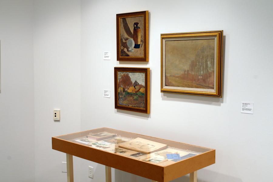 Three artworks on a wall. On the top left are two small black strips with brown shapes on them, and a small black square beneath them with a white letter A on it, all sharing a frame. On the bottom left is an abridged version of the poem Rhapsody on a Windy Night by T S Eliot written in black gothic script on a beige paper, and a stylized first letter T in yellow on blue. On the right is a painting of a black jug seen from above on a red and brown background.