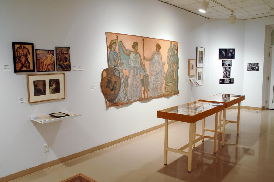 A gallery wall and two display cases of archival materials. From left to right, the wall includes: three artworks of a nude woman paired with two photographs of a clothed woman and a shelf of indiscernible pieces. Then a large painting imitating a Greco-Roman fresco featuring figures in togas, a red-figure vase, and a statue. Then three indiscernible works hung in frames, with another on a shelf. Then five indiscernible black and white photographs.
