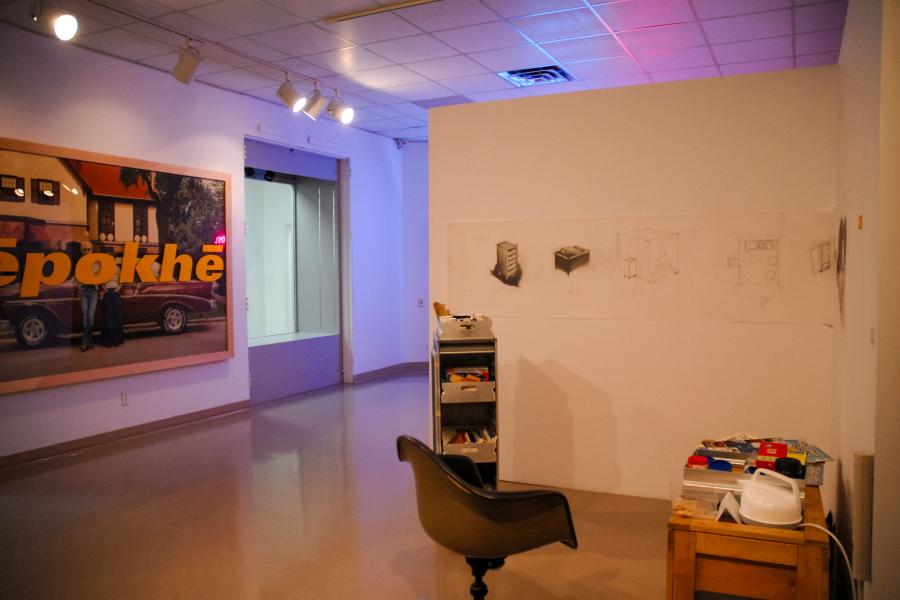 Installation view of a contemporary art exhibition featuring a workspace with a desk, chair, and rolling cabinet, charcoal drawings on partition walls, and a neon light installation illuminating the gallery in the background.
