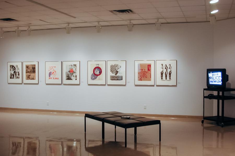 Installation view of the Peace Is Patriotic exhibition, with a row of framed anti-war prints displayed along a gallery wall, a bench in the foreground, and a CRT television showing a video installation.