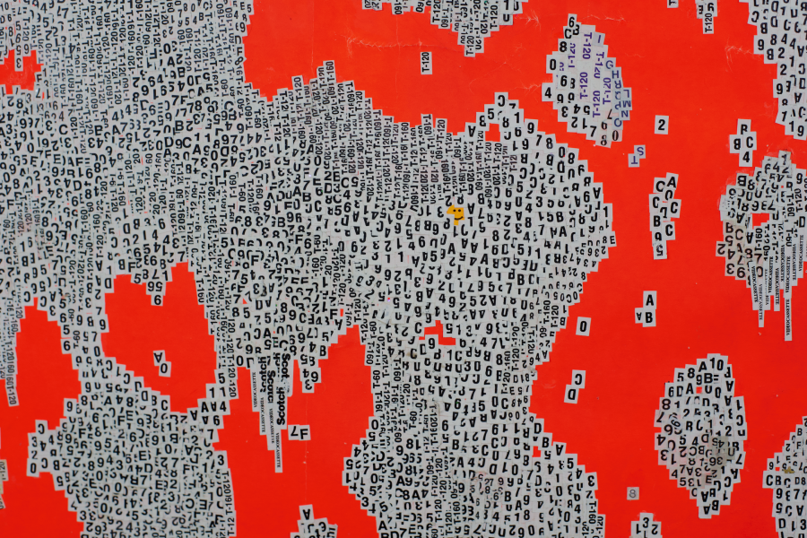 Detail of an artwork by Maya de Forest featuring a vivid red-orange background covered with dense, irregular clusters of cut-out letters, numbers and printed labels in black on white, arranged in fragmented, overlapping patterns.