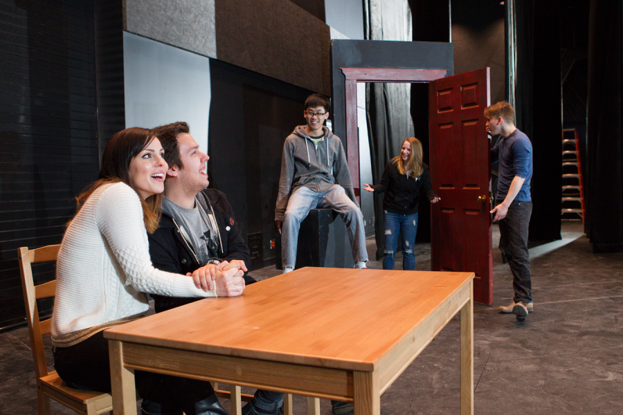 Five theatre students on a stage. Two sit at a table and one stands in a doorway surrounded by the other two.