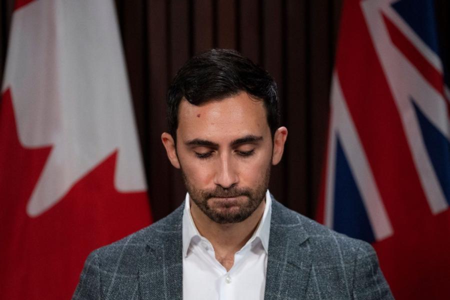Stephen Lecce, Minister of Education for Ontario, looking down in shame in front of Canadian and Ontario flags.