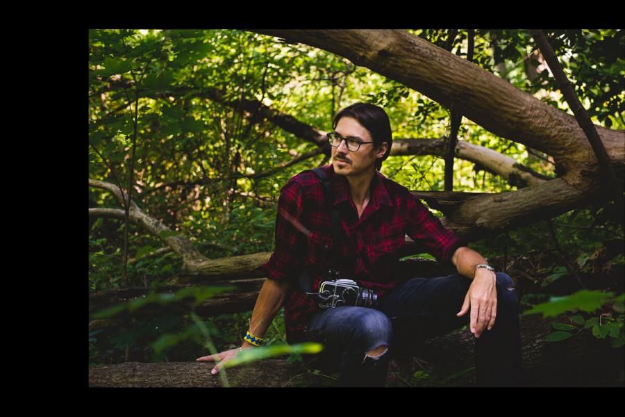 Photo of Aaron Vincent Elkaim sitting in a forrest with a camera.