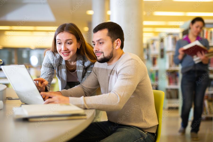 Male and female student looking at laptop screen, in a university library setting.