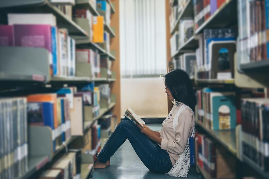 Woman sitting on the floor of a library in between two stacks of books, reading a book.
