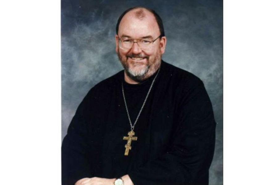 Headshot of priest in black cloak with large cross necklace around his neck.
