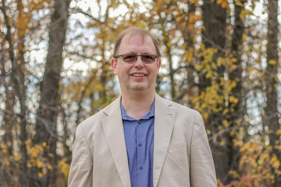 Man wearing glasses in front of block of trees with fall coloured leaves.