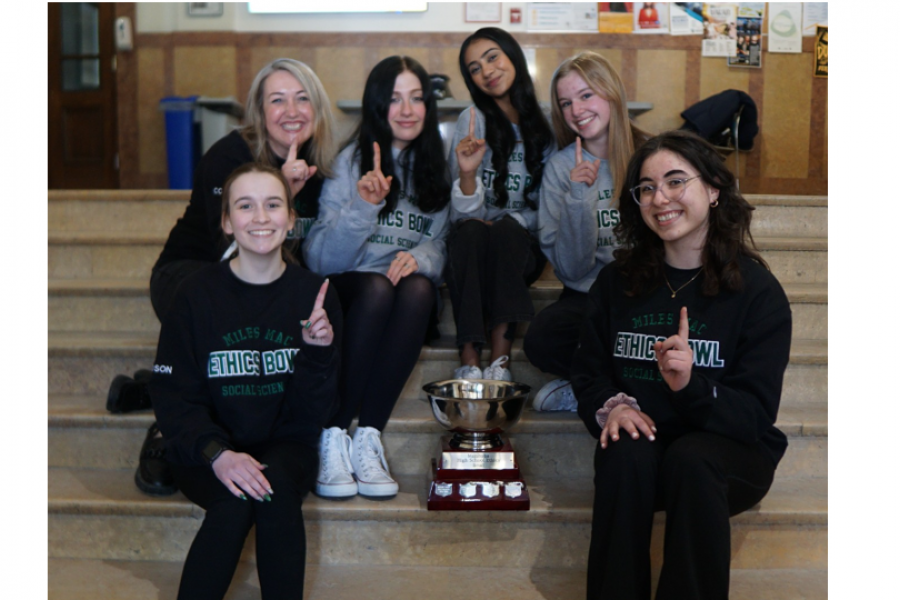 Five students and one teacher sitting around a trophy all holding up a winning finger.