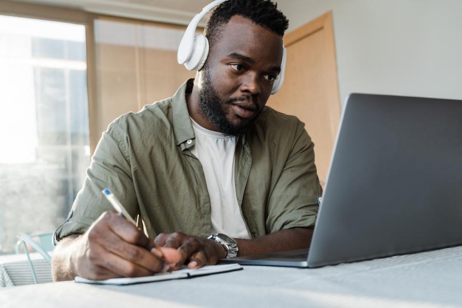 Student looking at laptop wearing white headphones and writing in a notebook.