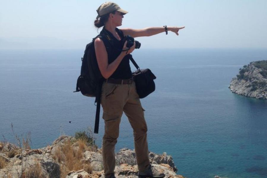 Person in hiking gear holding a camera standing on a cliff pointing out to sea.