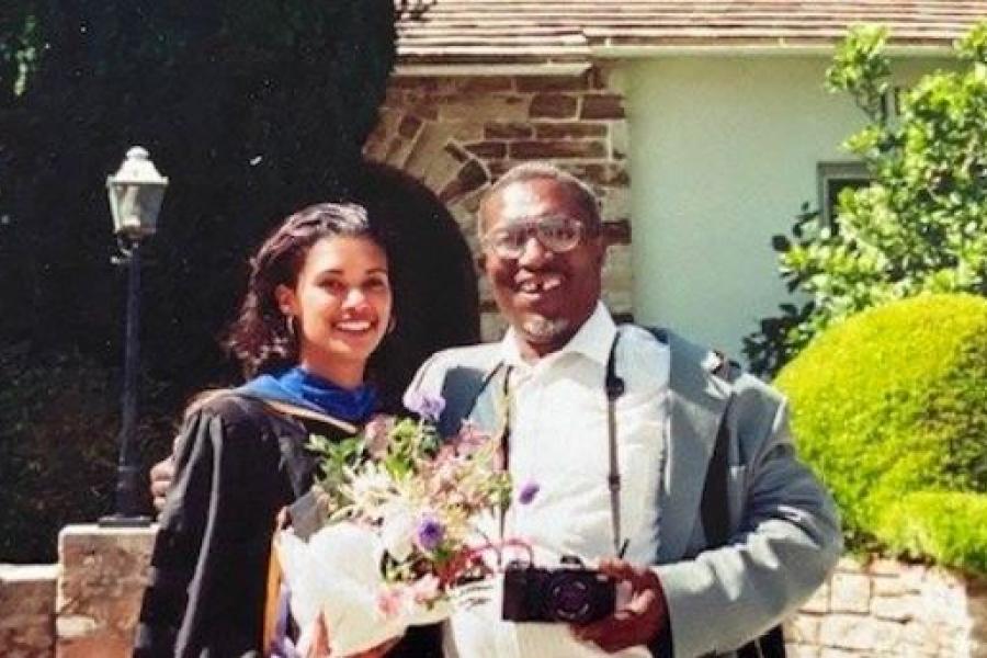 A woman in university graduation gowns stands with a man holding a camera.
