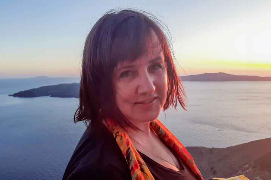 A headshot of Elena Baraban wearing a scarf, with a sunset and ocean background.