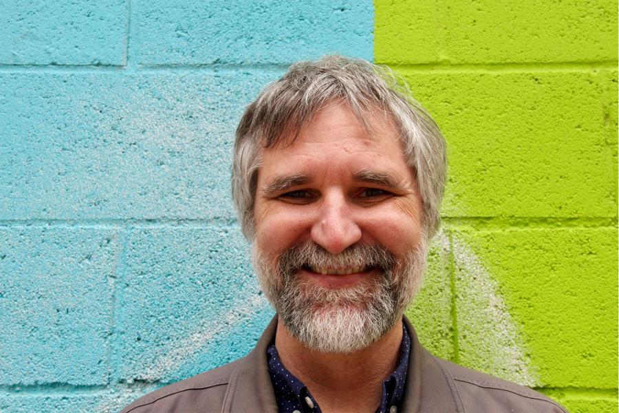A headshot of Greg Bak smiling in front of a colorful wall
