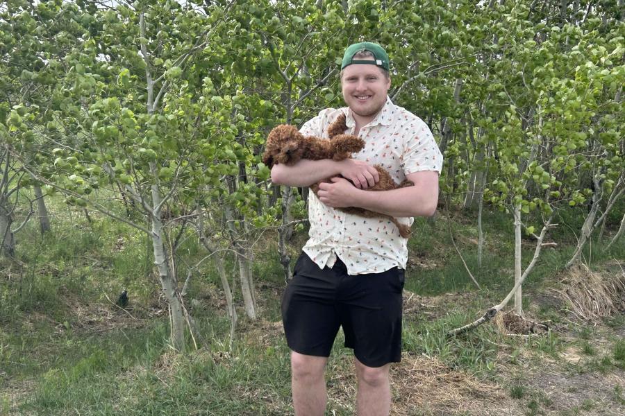Man standing in front a bunch of bushes, holding a small, brown dog in his arms.