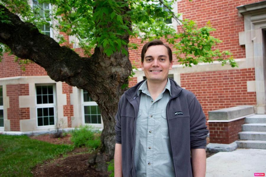 A person standing in front of a large tree with a brick building behind.