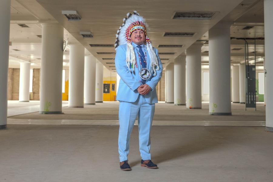 Man wearing a suit and traditional Indigenous headress stands in a large empty warehouse.