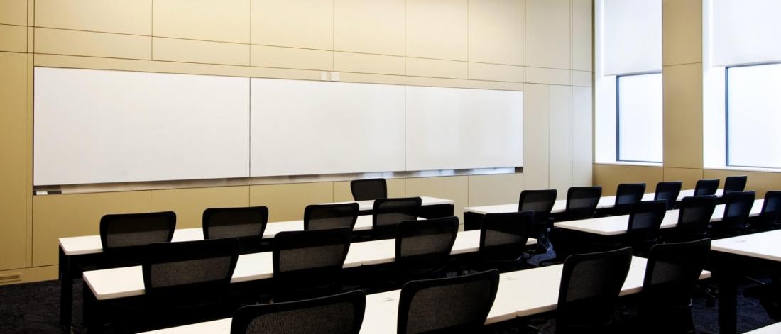 A large classroom with several narrow rows of tables and chairs and a large whiteboard at the front of the room.