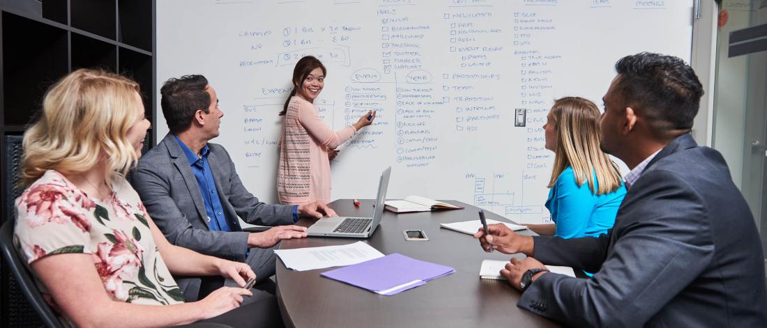 Four people seated around an office table watch as a woman writes a list on a whiteboard.