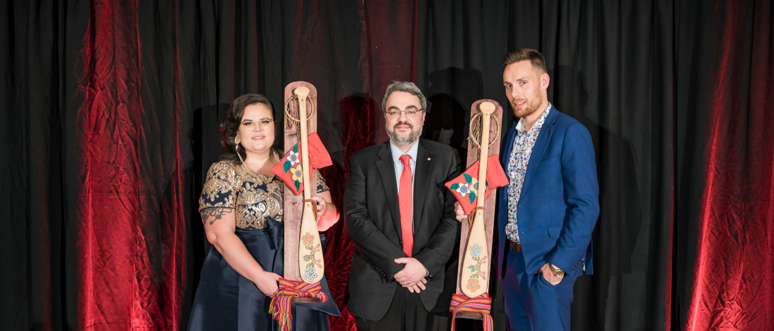 Three people dressed in business formal clothing, the people on the outside are holding an award that is a small paddle with decorative flowers engraved.