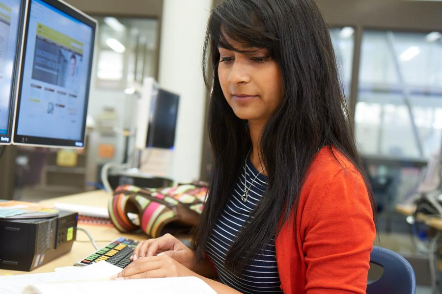 A student works intently seated at a computer work station while flipping through the pages of a book.