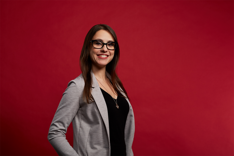 Woman with black shirt, grey blazer, and glasses smiling with a dark red, solid background.