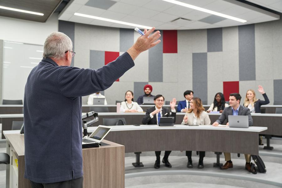 A male professor teaching a classroom full of students in the Asper School of Business Drake building. Some students have their hand up and are wearing business casual clothes.