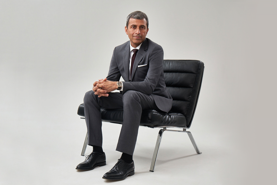 Ash Modha wearing a grey suit sitting on a black leather lounge chair. He is looking at the camera.
