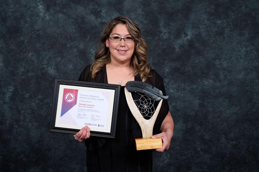Woman holding framed certificate and award that looks like a dreamcatcher on an antler. She is wearing a dress and smiling.